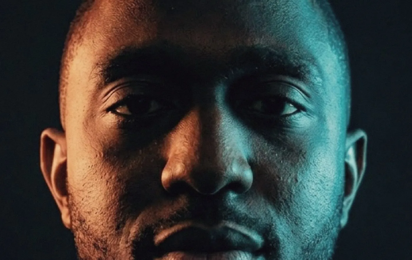 A man's face in tight close-up against black, lit from both sides at once: warm amber on the left, cold cyan on the right, the two meeting down the line of his nose. Brow to chin fills the frame and he looks straight at the camera.