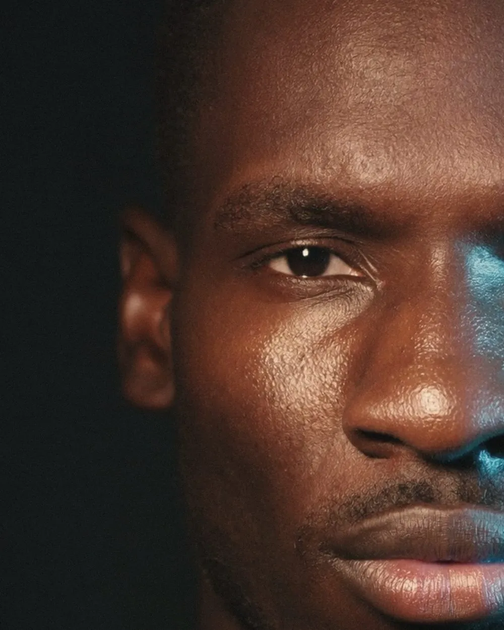 A tall crop holding one half of a man's face at the right of the frame, the rest black. Warm amber light falls across the brow, one open brown eye, the cheek and the lips, with a thin cold blue highlight along the far edge of the nose and mouth.