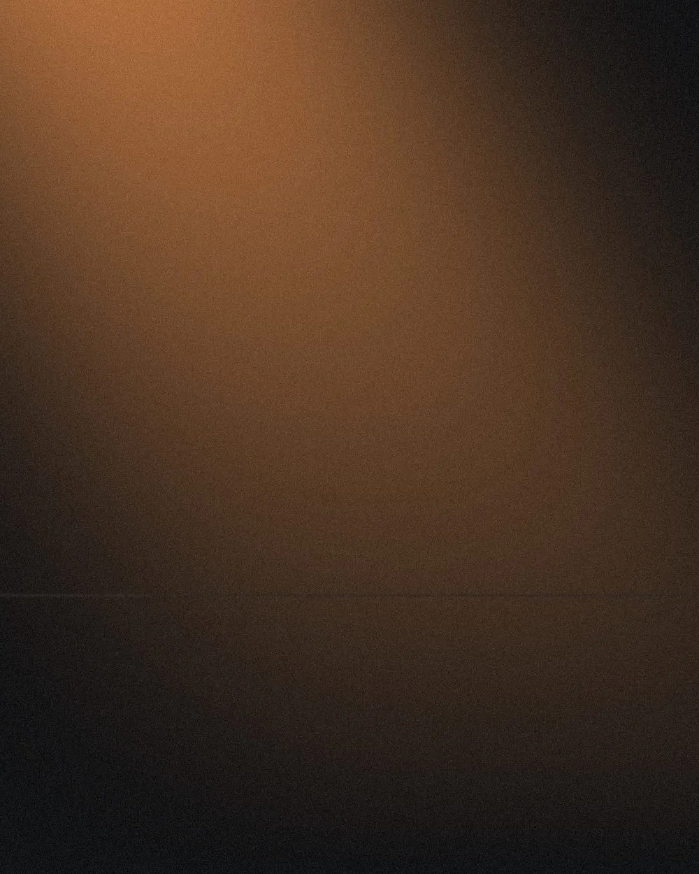 An empty studio corner washed in soft amber light, brightest at the top left and falling away to black at the right and along the bottom, with a faint seam where the wall meets the floor.