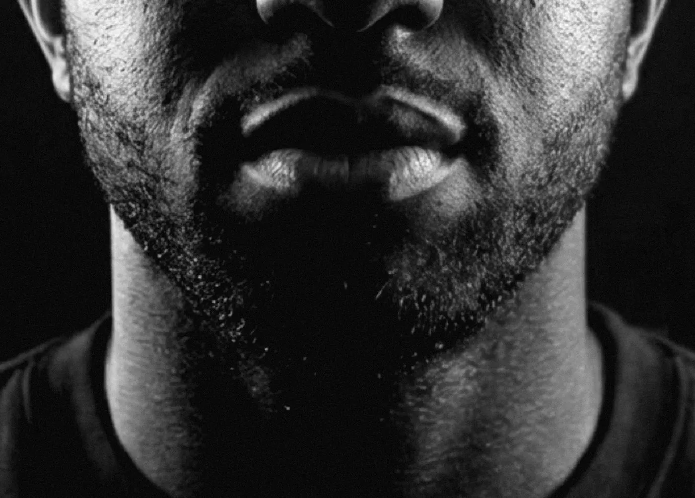 A black and white crop of the lower half of a man's face, seen head-on: nostrils at the top edge, closed lips, stubble across the jaw and neck, and the collar of a dark t-shirt at the bottom. Hard side light leaves the centre in deep shadow.
