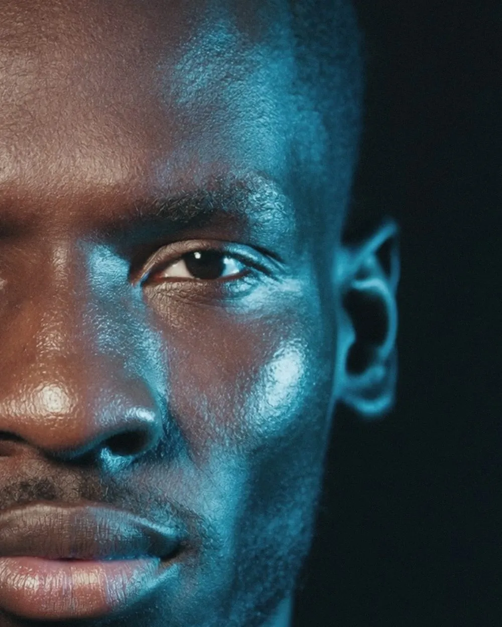 A tall crop of one side of a man's face against black. Warm light holds the near cheek, nose and lips at the left edge, while a strong teal light wraps the brow, the open eye, the cheekbone and the ear.