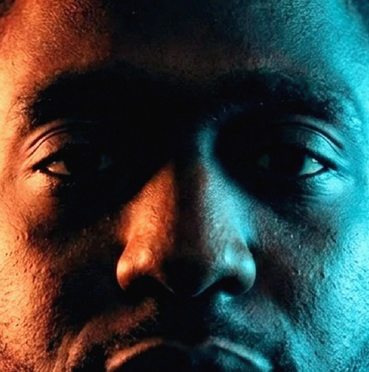 A near-square crop across both of a man's eyes and the bridge of his nose. Orange light covers the left half of the face and cyan the right, the two colours meeting along the nose, his gaze slightly raised.