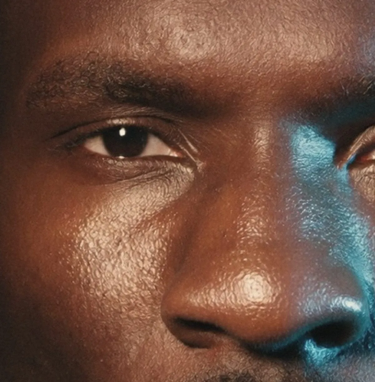 A close crop of one eye and the near side of a man's nose in warm brown light, a small white catchlight in the iris, while a cold blue-green light strikes the far cheek and the far edge of the nose at the right of the frame.
