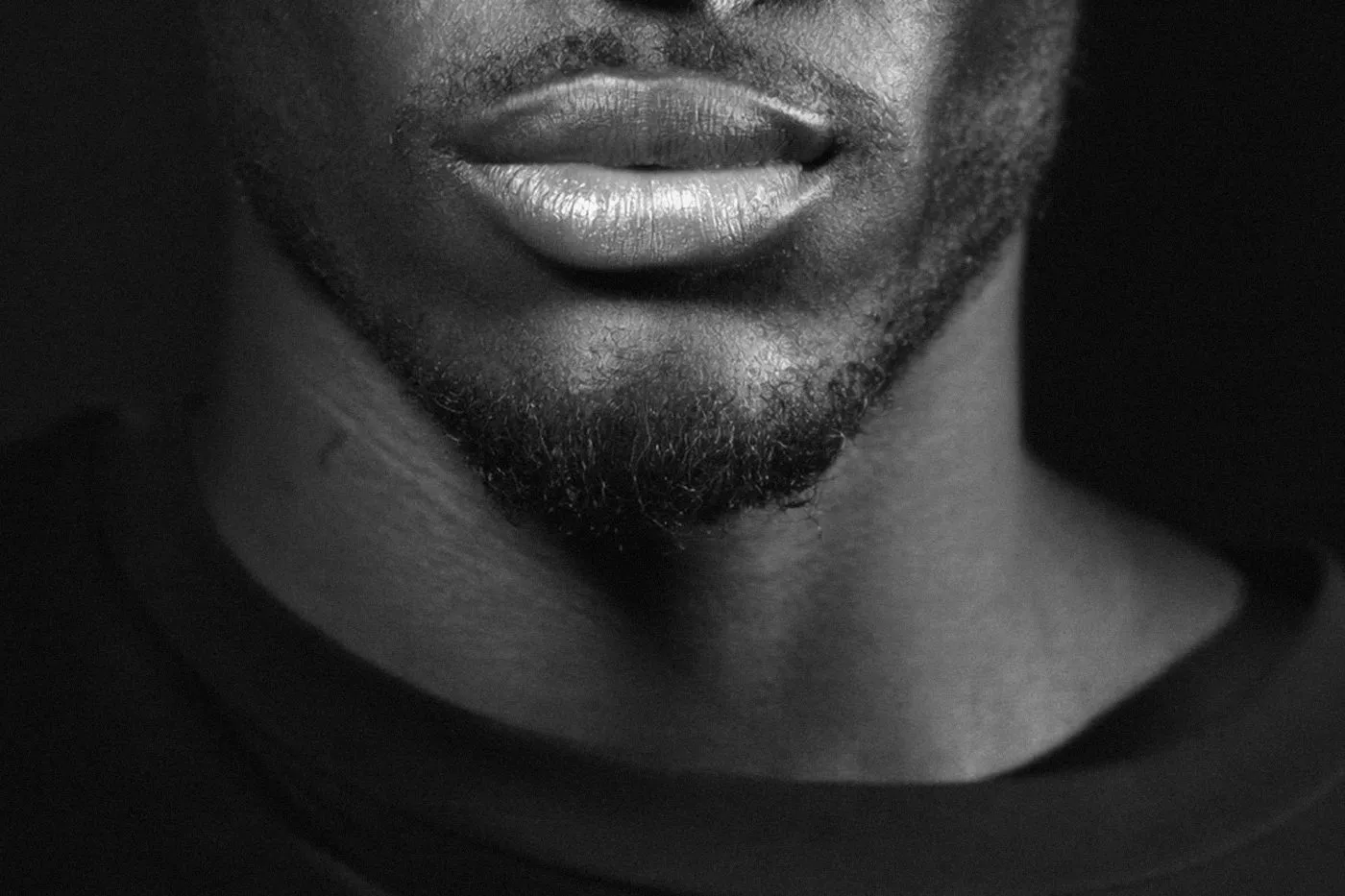 A black and white crop from the nose down: parted lips, a short beard along the chin and jaw, the neck, and a dark crew collar. The light comes from the left and the right of the frame falls away into black.